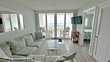 A living room with a sofa, a coffee table, and a TV. There is a balcony with a view of the ocean.