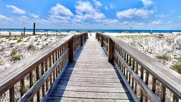 Ein hölzerner Steg führt zu einem Strand mit weißem Sand und einem klaren, blauen Himmel.