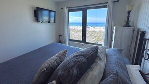 Ein Schlafzimmer mit einem Bett, einem an der Wand montierten Fernseher und einem Fenster mit Blick auf den Strand.
