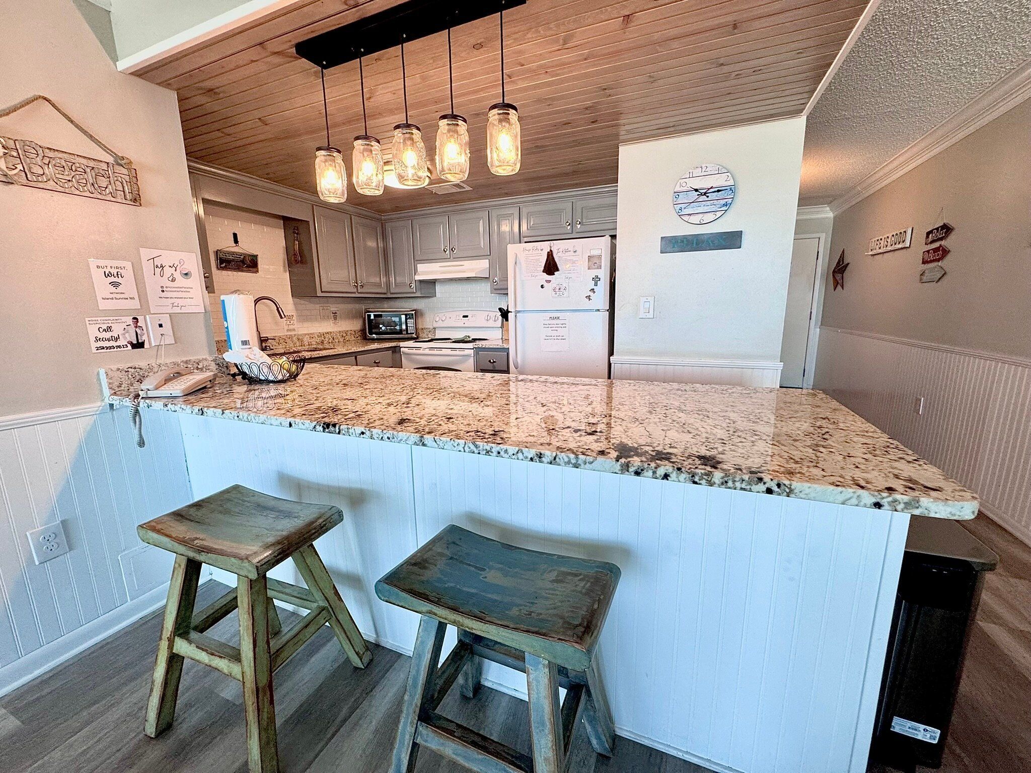 A kitchen with a granite countertop, wooden stools, hanging pendant lights, and a refrigerator with magnets.