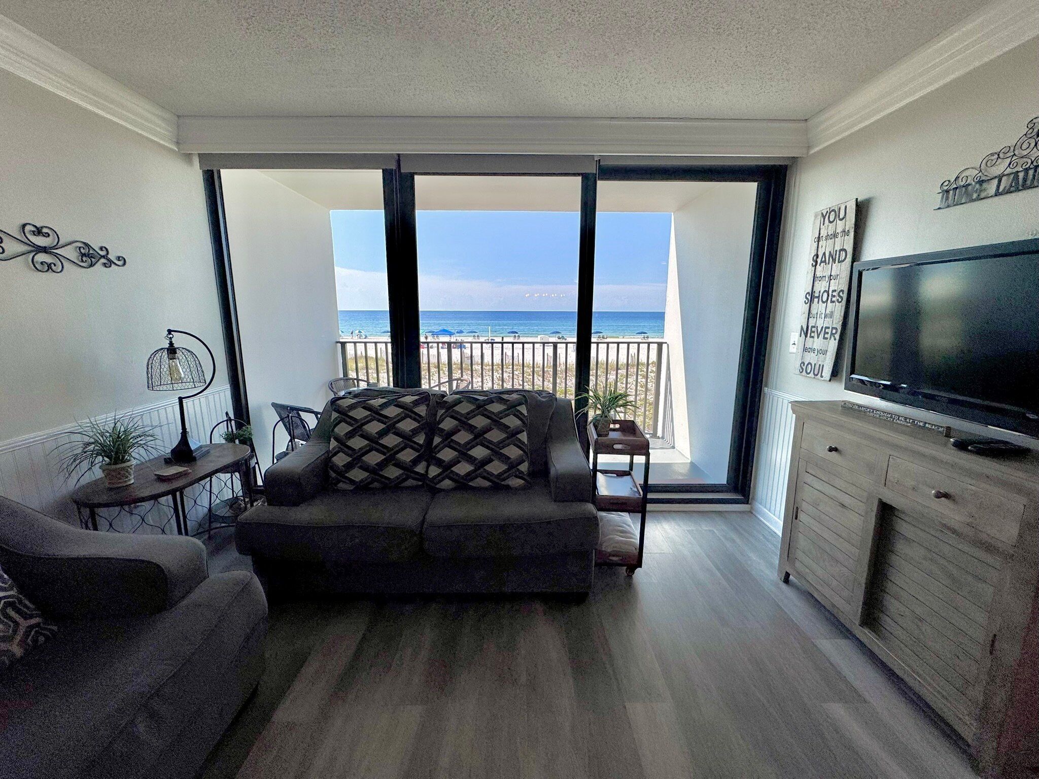 A living room with a sofa, a TV, and a balcony with a view of the beach.