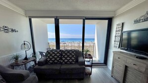 Ein Wohnzimmer mit einer Couch, einem Fernseher und einem Balkon mit Blick auf den Strand.