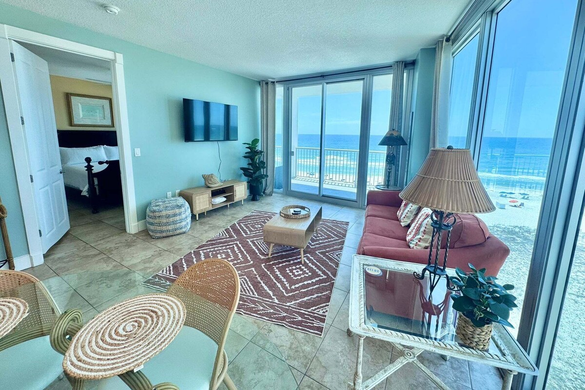 A spacious living area with a large window overlooking the beach, a flat-screen TV mounted on the wall, and a cozy seating arrangement.