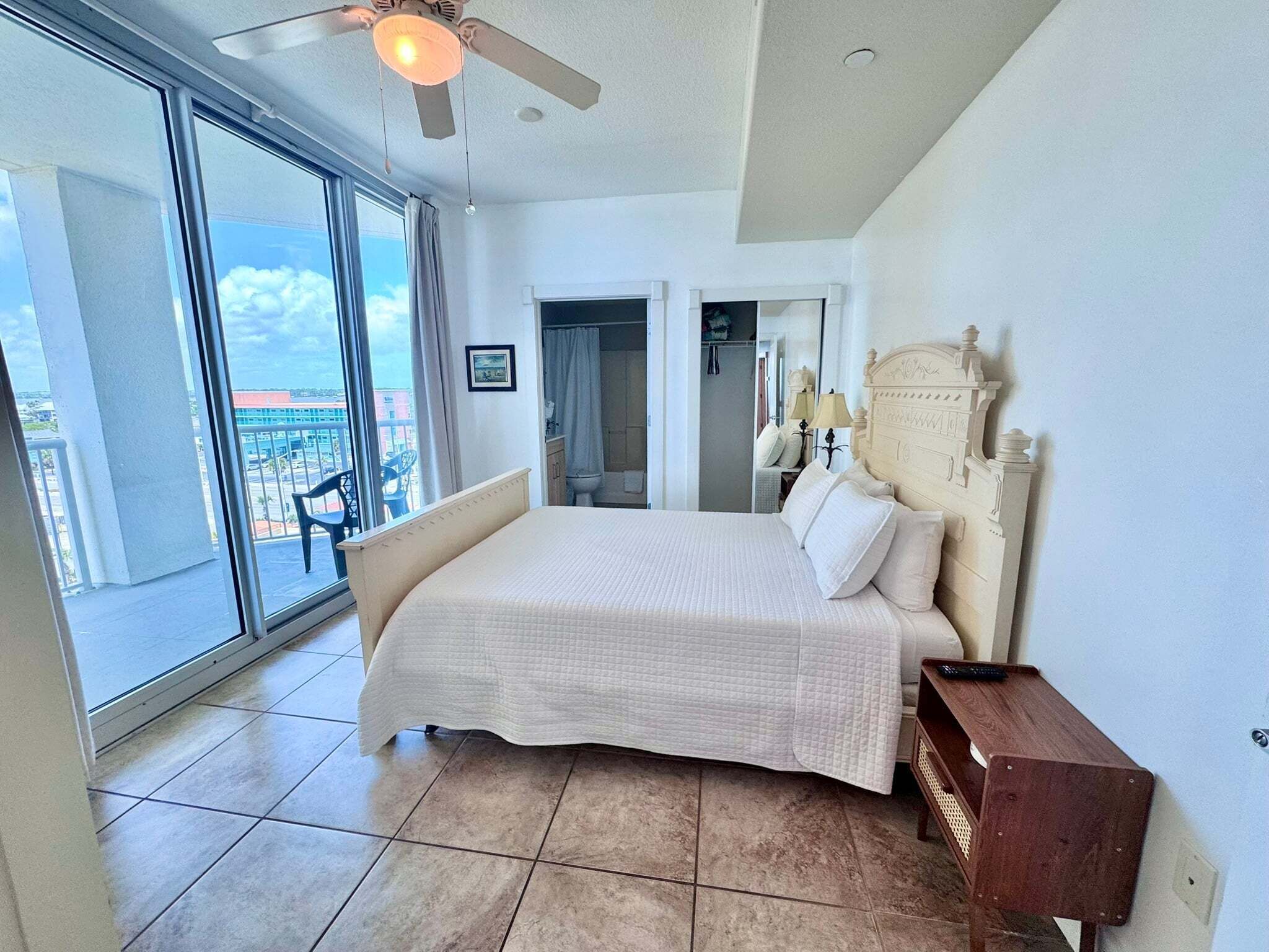 A bedroom with a large bed, a ceiling fan, a nightstand, and a view of the outside.