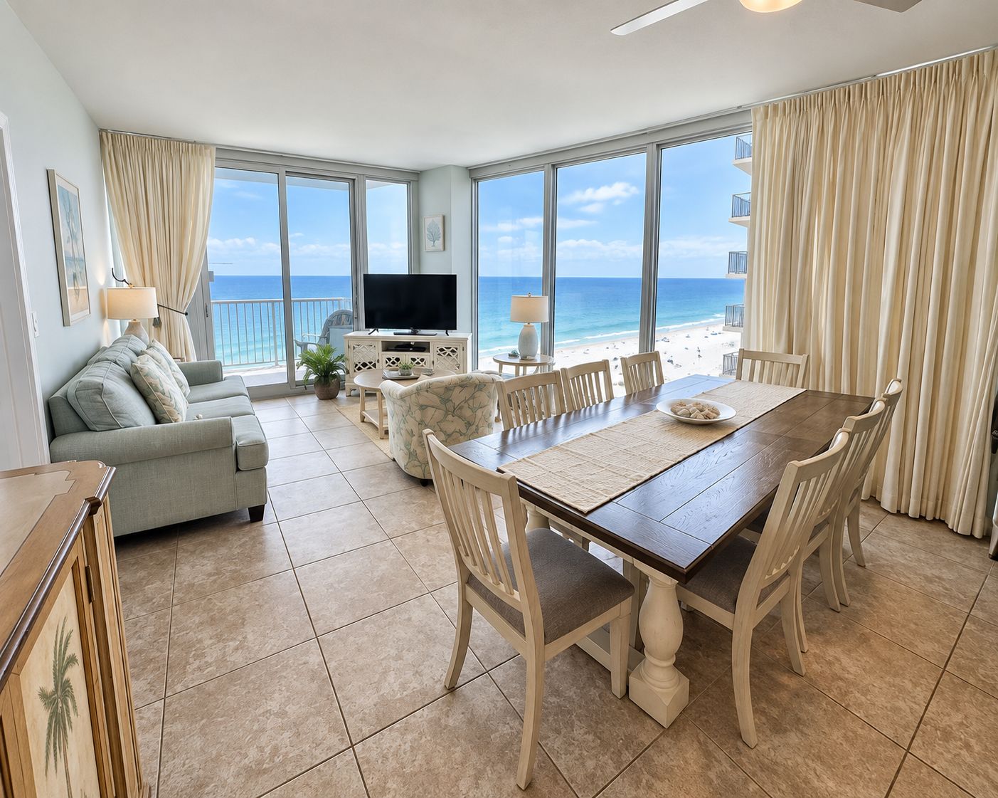 A spacious living area with a large window overlooking the beach, a dining table with chairs, a sofa, and a television.