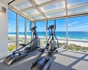 A gym with two stationary bikes and a large window overlooking a beach. - Island Tower (Gulf Shores)
