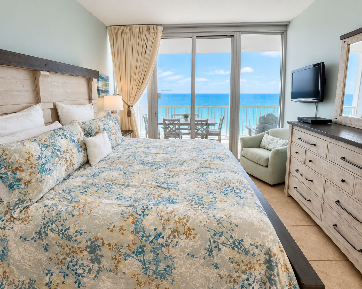 A bedroom with a large bed, a television, a dresser, and a balcony with a view of the ocean.