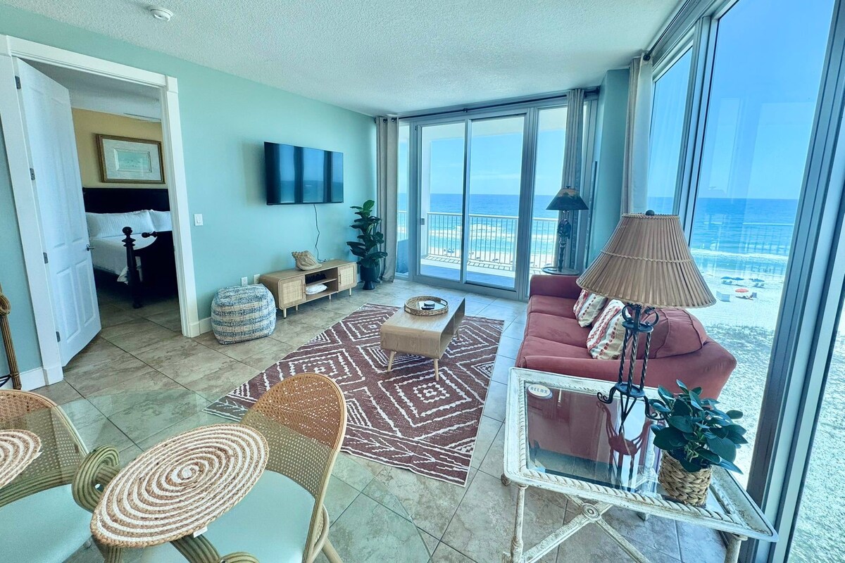 A spacious living area with a large window overlooking the beach, a flat-screen TV mounted on the wall, and a cozy seating arrangement.