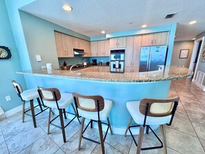 A kitchen with a central island, bar stools, wooden cabinets, stainless steel appliances, and a light blue wall. - Island Tower (Gulf Shores)