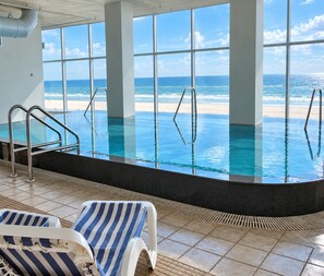 An indoor swimming pool with clear ocean views, tiled flooring, and striped lounge chairs. - Island Tower (Gulf Shores)