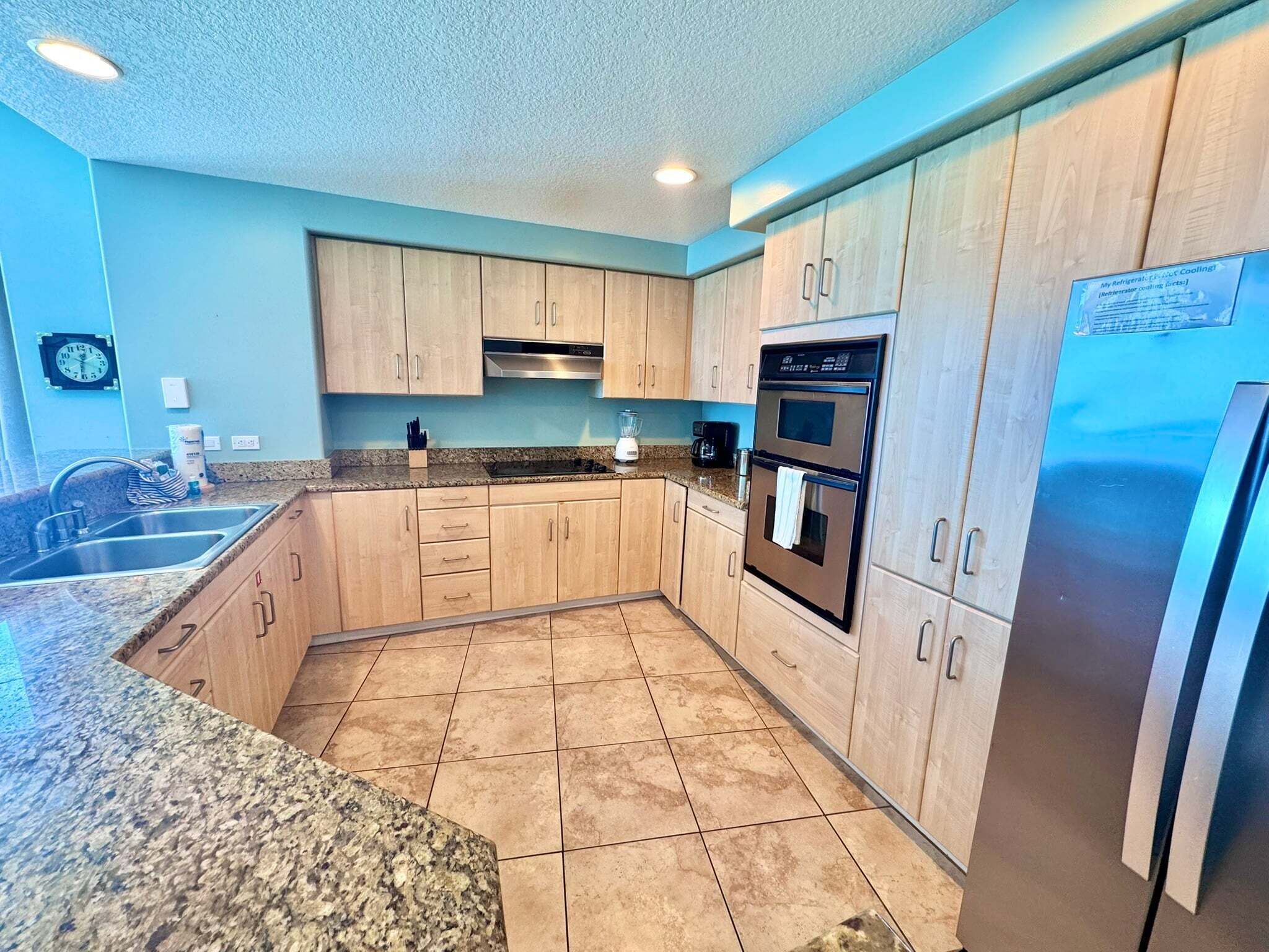 A kitchen with light wood cabinets, a stainless steel refrigerator, a double oven, and a granite countertop.