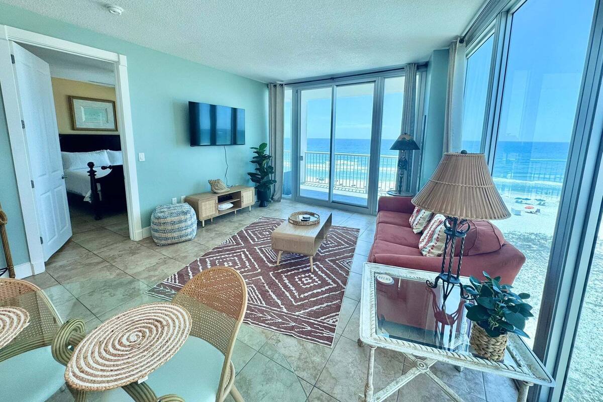 A spacious living area with a large window overlooking the beach, a flat-screen TV mounted on the wall, and a cozy seating arrangement.