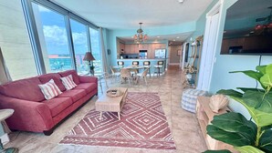 A spacious living area with a red sofa, a dining table, and chairs, a kitchen in the background, and large windows offering a view. - Island Tower (Gulf Shores)