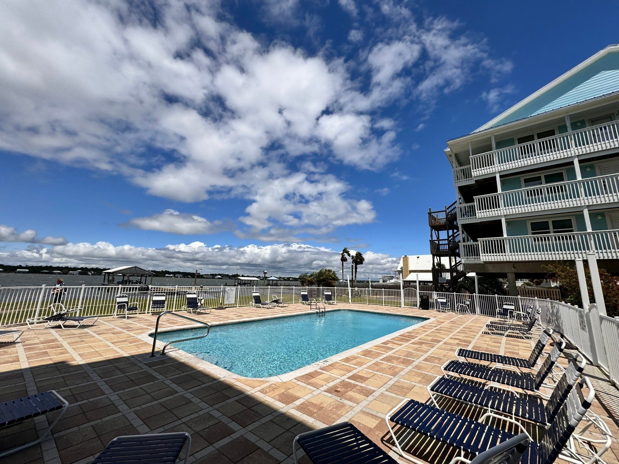 Un área de piscina con sillones de descanso, un cielo azul despejado y un edificio con balcones.
