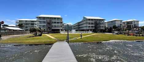 A waterfront property with a wooden dock leading to a grassy area and a building with multiple balconies.