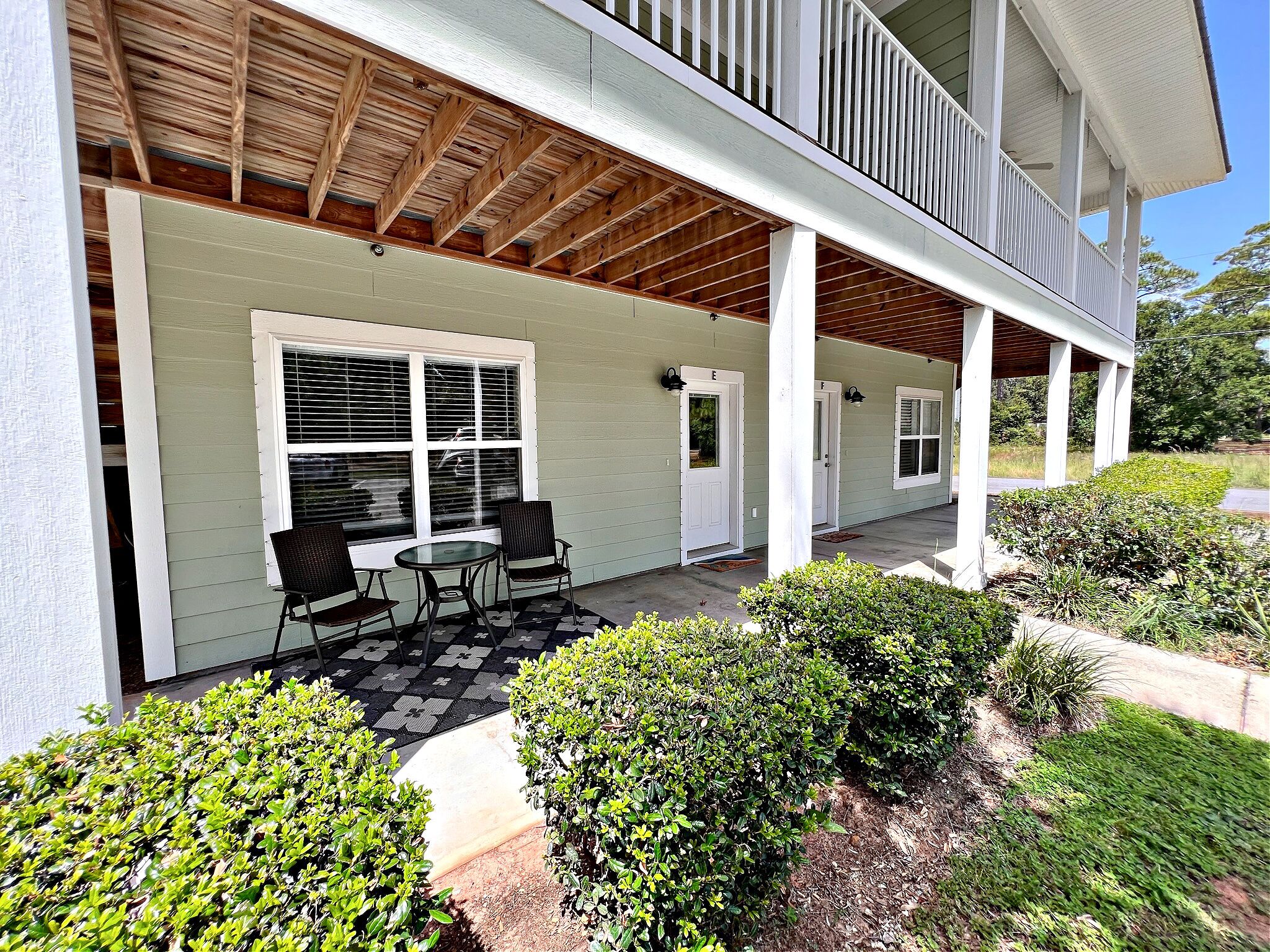 A covered outdoor seating area with a table and chairs, surrounded by well-maintained bushes and a paved walkway.