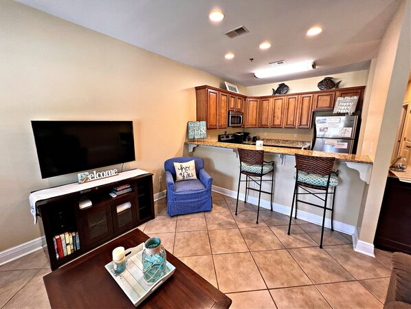 A compact kitchen with wooden cabinets, a built-in microwave, and a refrigerator. There is a bar area with two stools and a television in the living area. - Laguna Breeze (Gulf Shores)