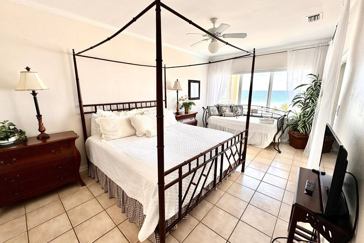 A four-poster bed with a canopy, a desk with a chair, a television, and a view of the ocean.
