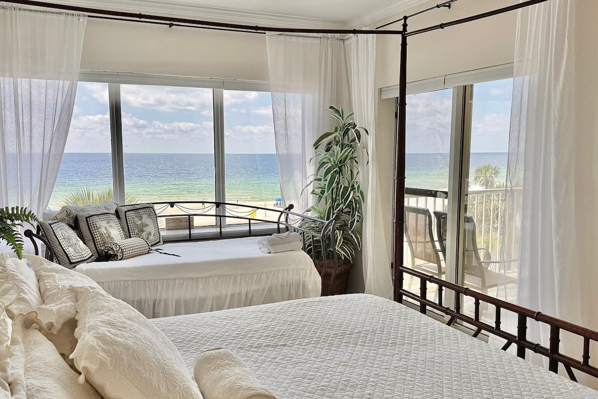 A bedroom with a large bed, a balcony with a view of the ocean, and a potted plant.