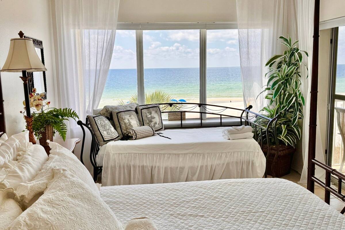 A bedroom with a bed, a nightstand, a lamp, and a view of the ocean.