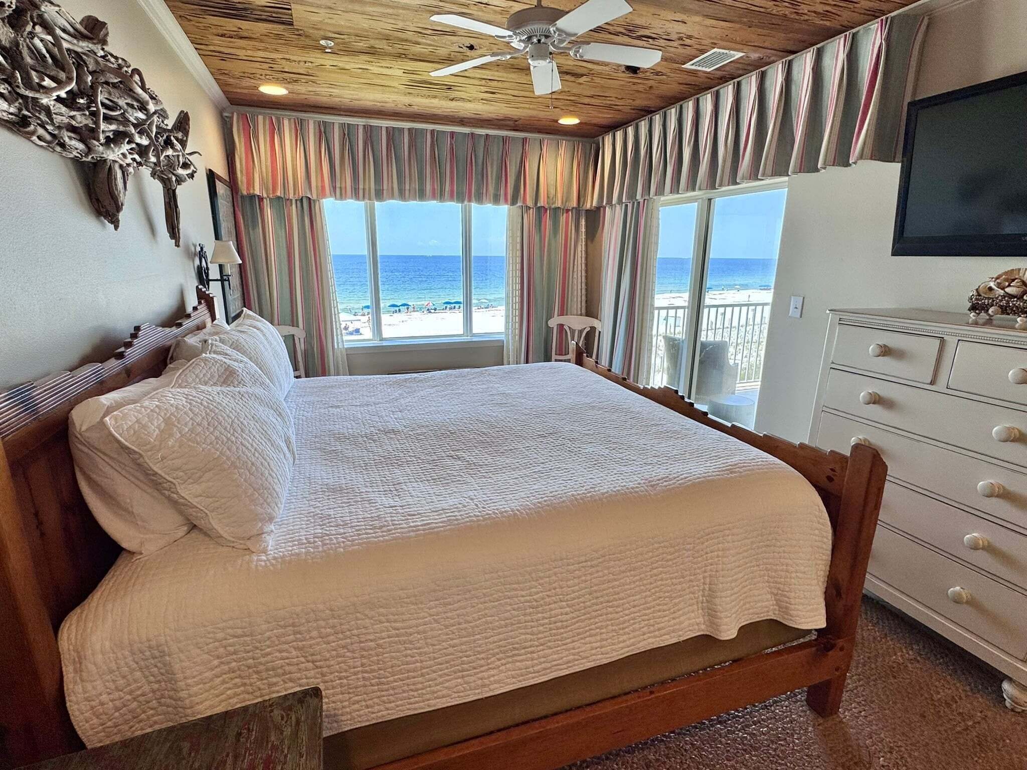 A bedroom with a large bed, a ceiling fan, a dresser, and a view of the beach.