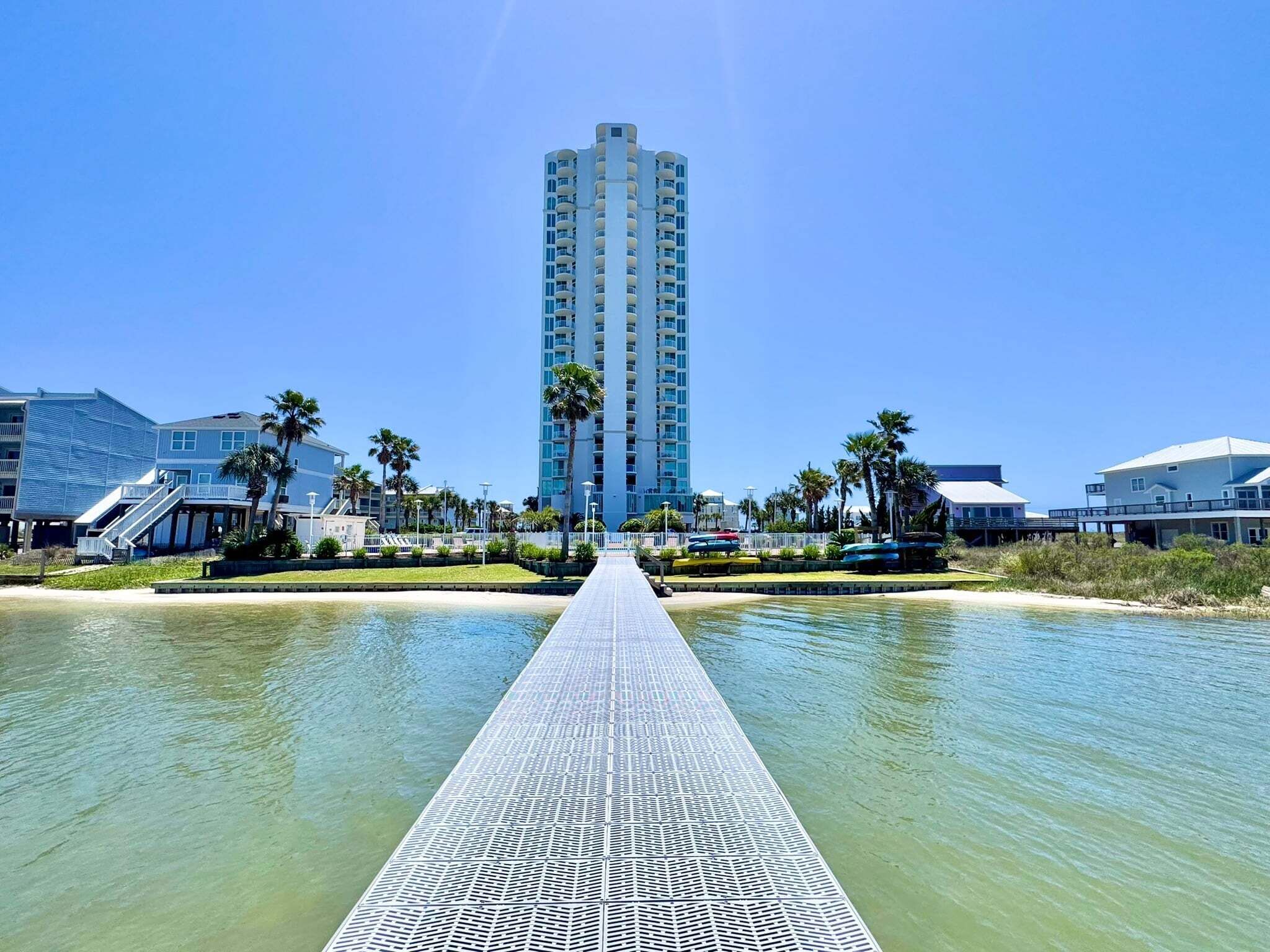 A waterfront hotel with a long pier leading to the beach, a tall building in the background, and palm trees.