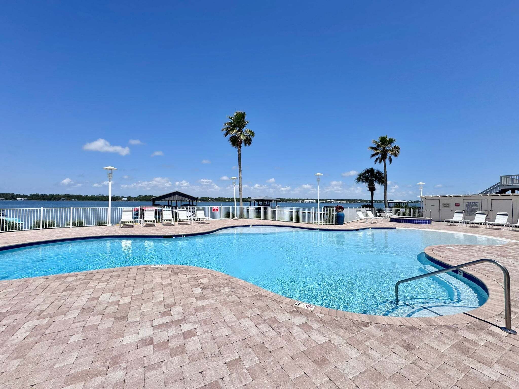 A swimming pool with a brick deck, lounge chairs, palm trees, and a view of the water.