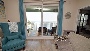 Un salon avec un fauteuil turquoise, un canapé beige et une vue sur la mer, visible à travers une porte-fenêtre coulissante.