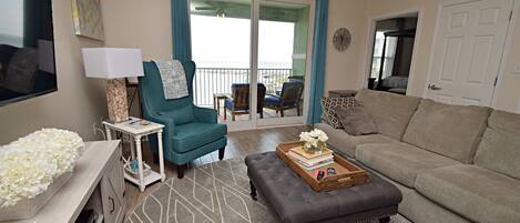 Un salon comprenant un canapé, un fauteuil turquoise, une table basse avec des livres et des fleurs, un téléviseur et un balcon avec des rideaux bleus.