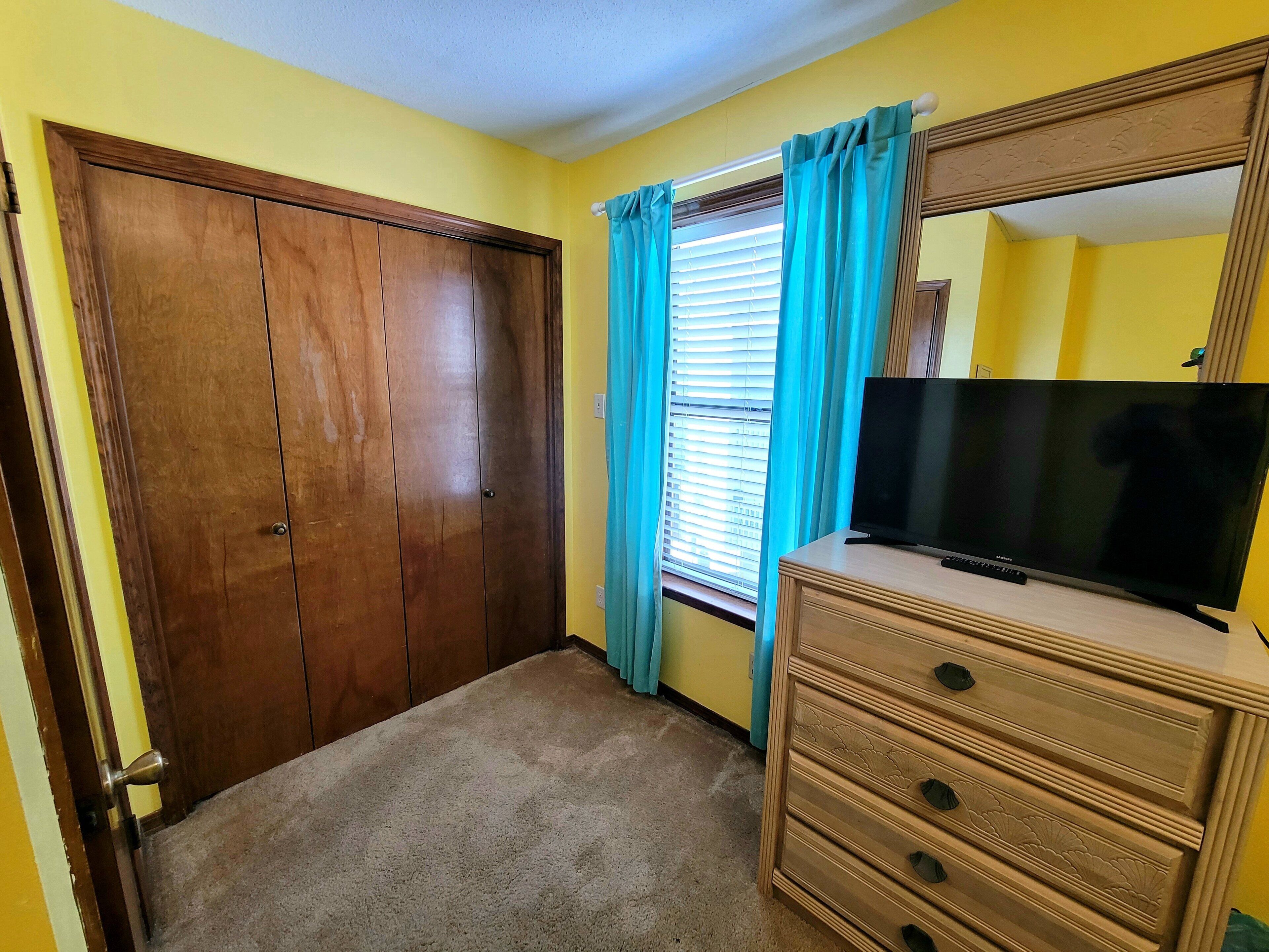 A room with a wooden wardrobe, a television on a dresser, and a window with blue curtains.