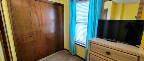 A room with a wooden wardrobe, a television on a dresser, and a window with blue curtains.
