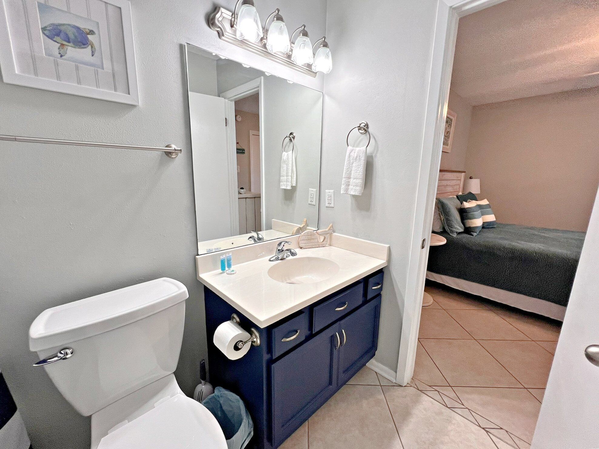 Un baño con inodoro, lavamanos y espejo. Hay un perchero con toallas y un cuadro enmarcado en la pared. La habitación tiene una puerta que da a otra habitación con una cama.