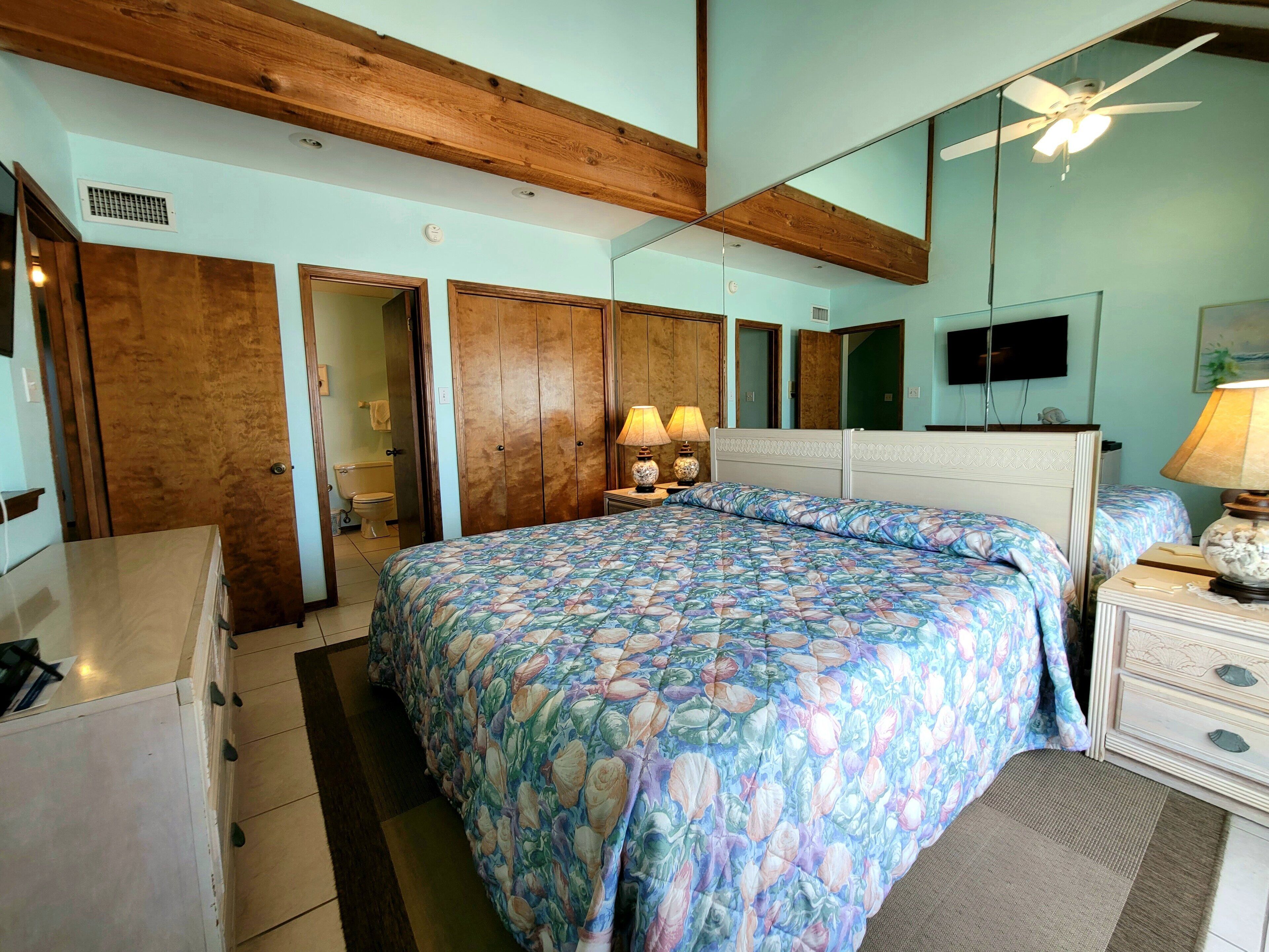 A bedroom with a large bed, wooden ceiling beams, a ceiling fan, a TV, and a bathroom visible through an open door.