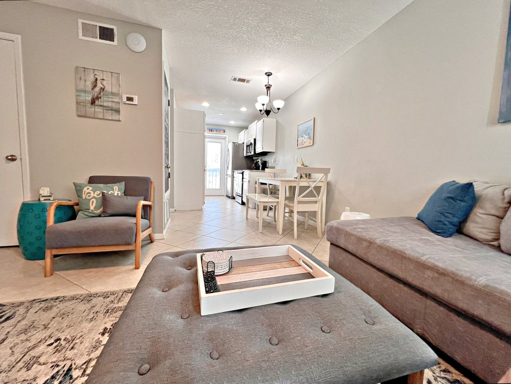 Un salon moderne avec un canapé gris, un pouf assorti, une table d’appoint de couleur turquoise et un coin repas comprenant une table et des chaises blanches.