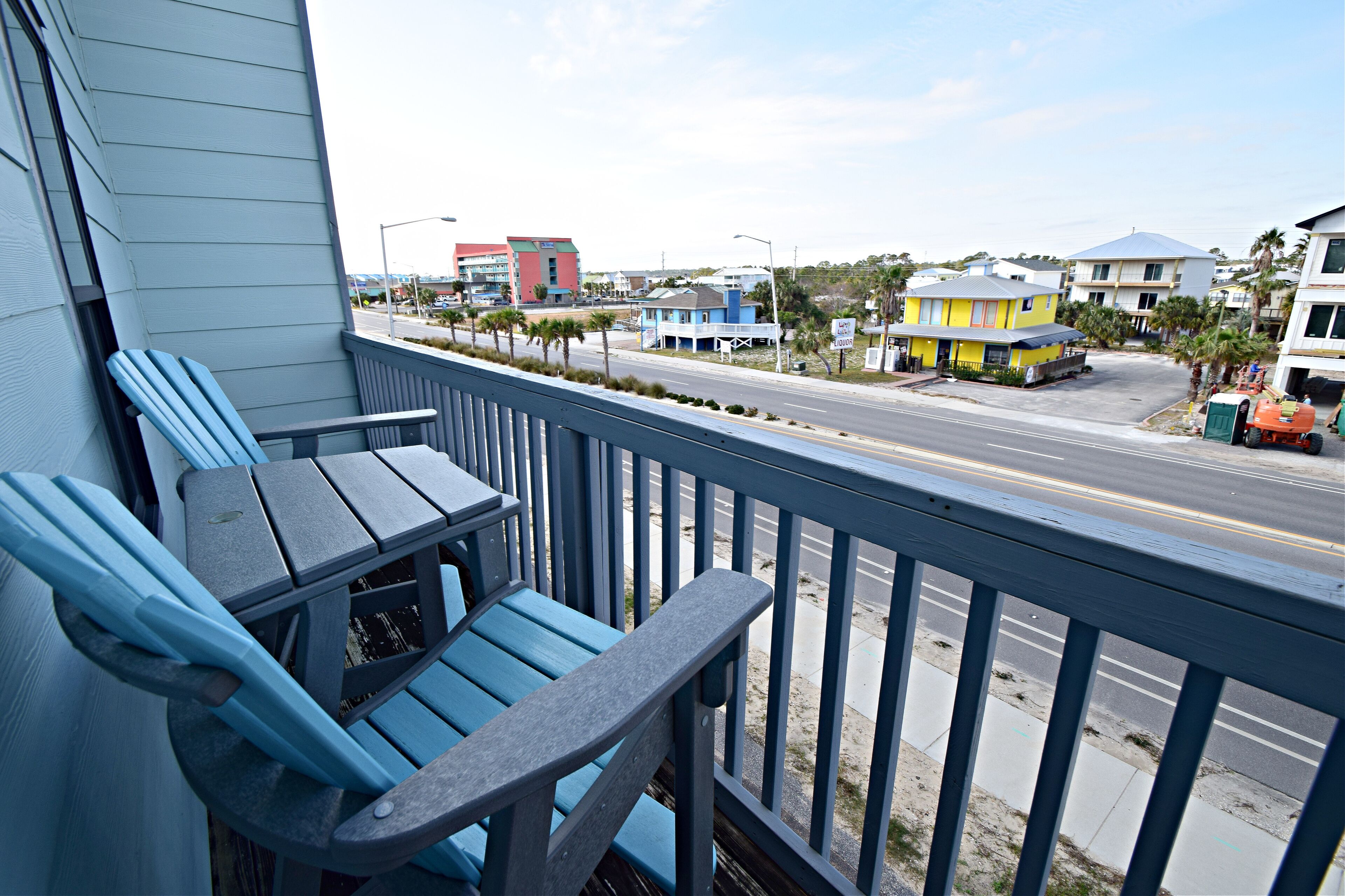 Een balkon met blauwe Adirondack-stoelen en een tafel, met uitzicht op een straat met gebouwen en een heldere lucht.