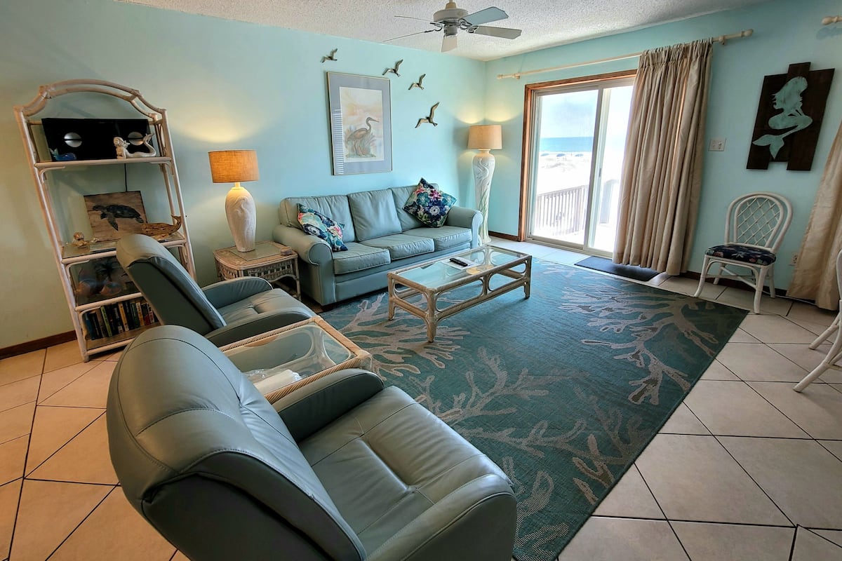 A living room with a sofa, armchairs, a coffee table, a bookshelf, and a view of the outdoors through a sliding door.