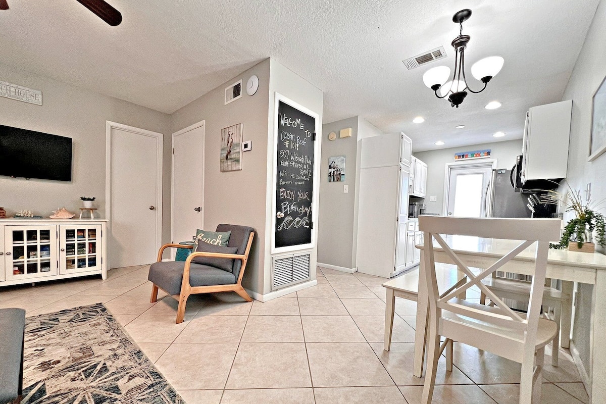 A spacious living area with a dining table, a kitchen in the background, and a large TV on the wall.