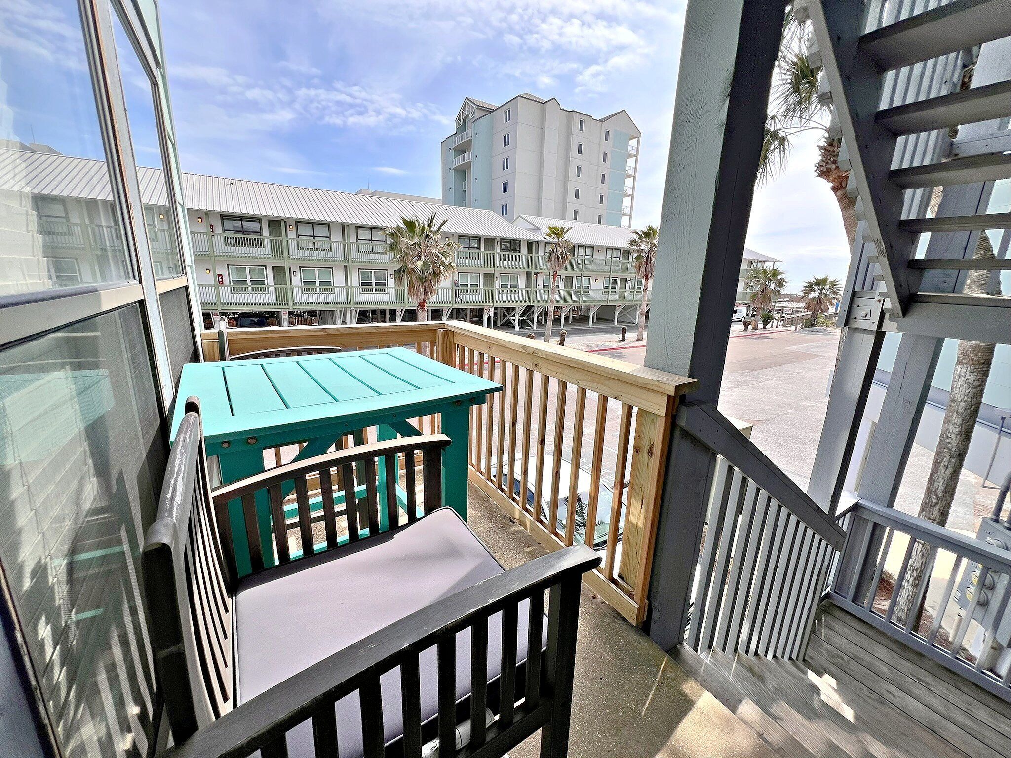 A balcony with a wooden table and chairs, a view of a multi-story building, and a clear sky.