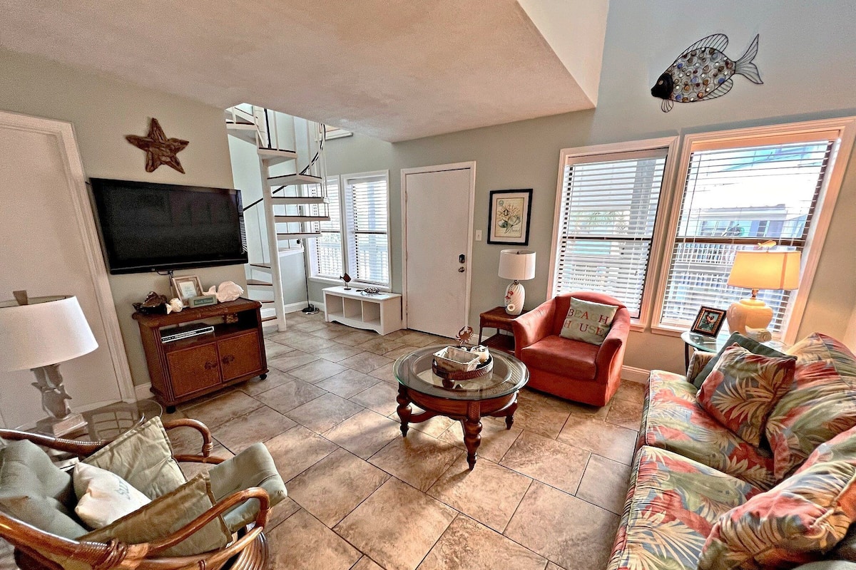 A living room with a TV, a red armchair, a wicker chair, a glass coffee table, and a colorful sofa.