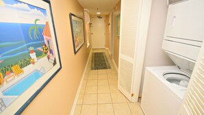A narrow hallway with a tiled floor leading to a door. There are framed pictures on the walls, a washing machine, and a door with a towel hanging on it. - Phoenix East (Orange Beach)