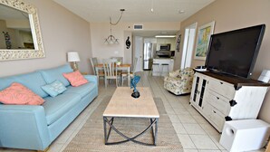 A living room with a blue sofa, a coffee table, a TV, and a kitchen in the background. - Phoenix East (Orange Beach)