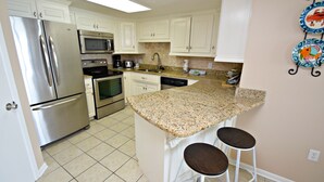 A kitchen with white cabinets, stainless steel appliances, a granite countertop, and two bar stools. - Phoenix East (Orange Beach)