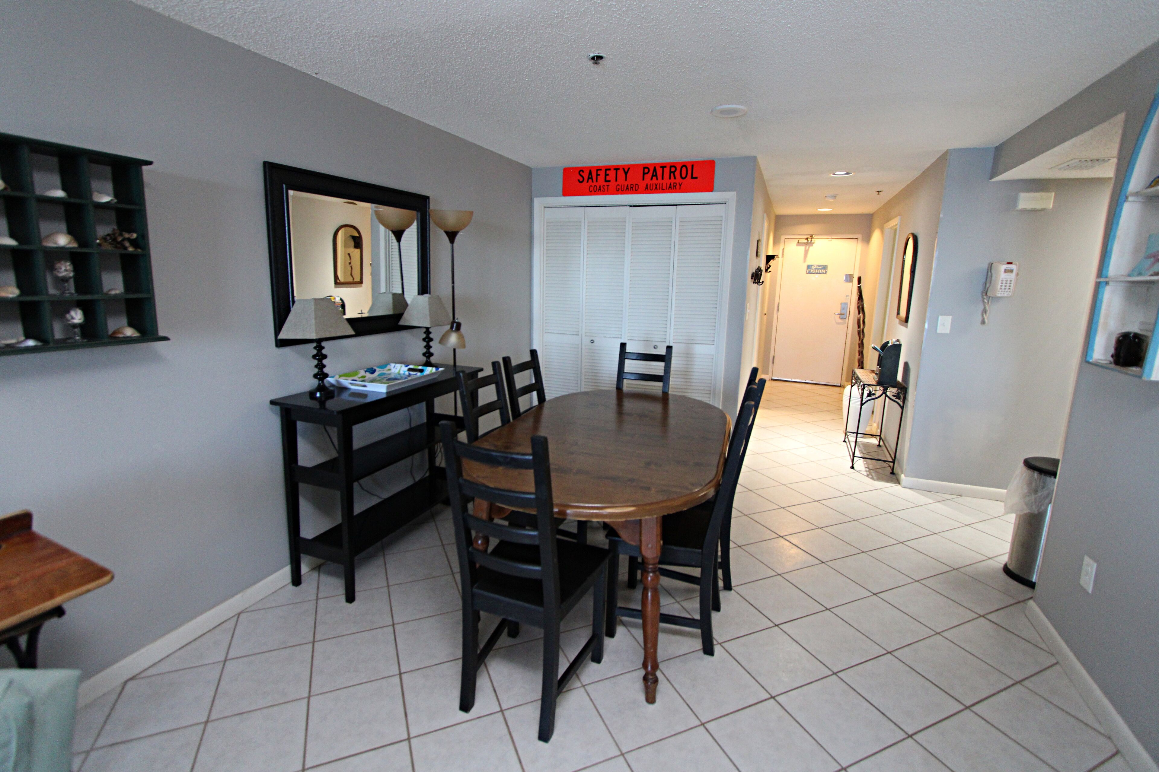A dining area with a round table and chairs, a mirror, a shelf with decorative items, and a sign reading "SAFETY PATROL COAST GUARD AUXILIARY".