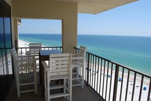 Ein Balkon mit weißen Stühlen und einem Tisch, der Blick auf einen Strand mit klarem, blauem Wasser und sandigem Ufer bietet.