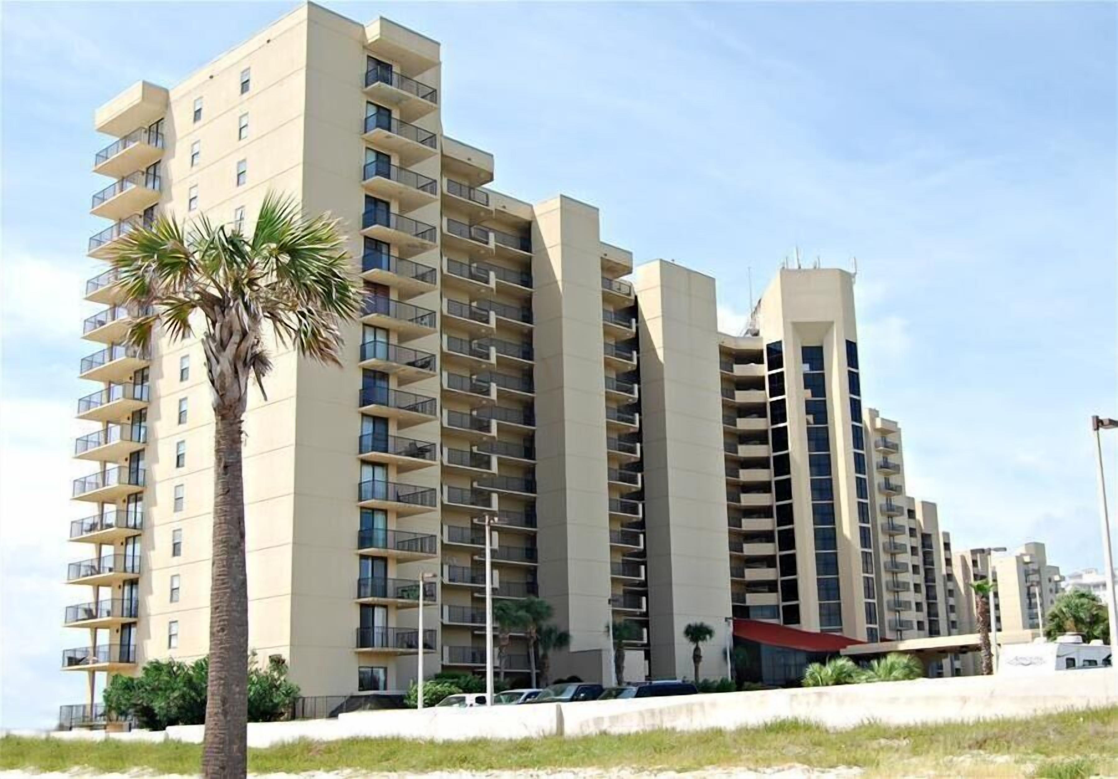 A multi-story hotel with balconies, a palm tree, and a parking area.