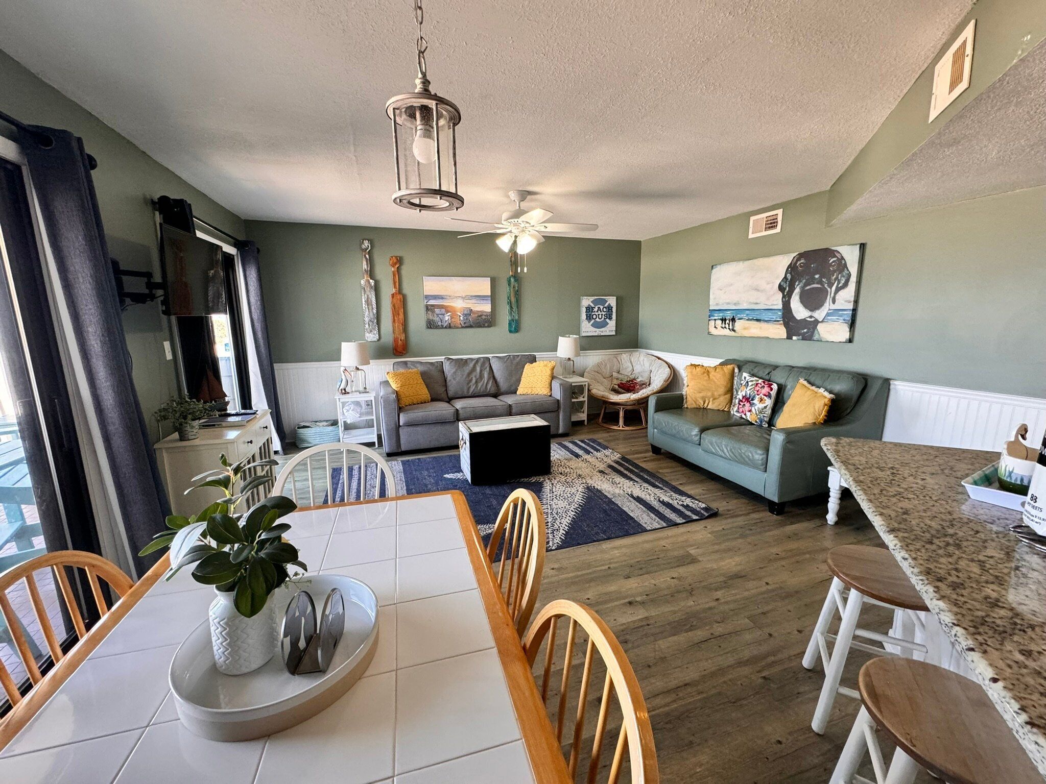 A modern living room with a grey sofa, a dining table with chairs, and a kitchen area with a granite countertop.