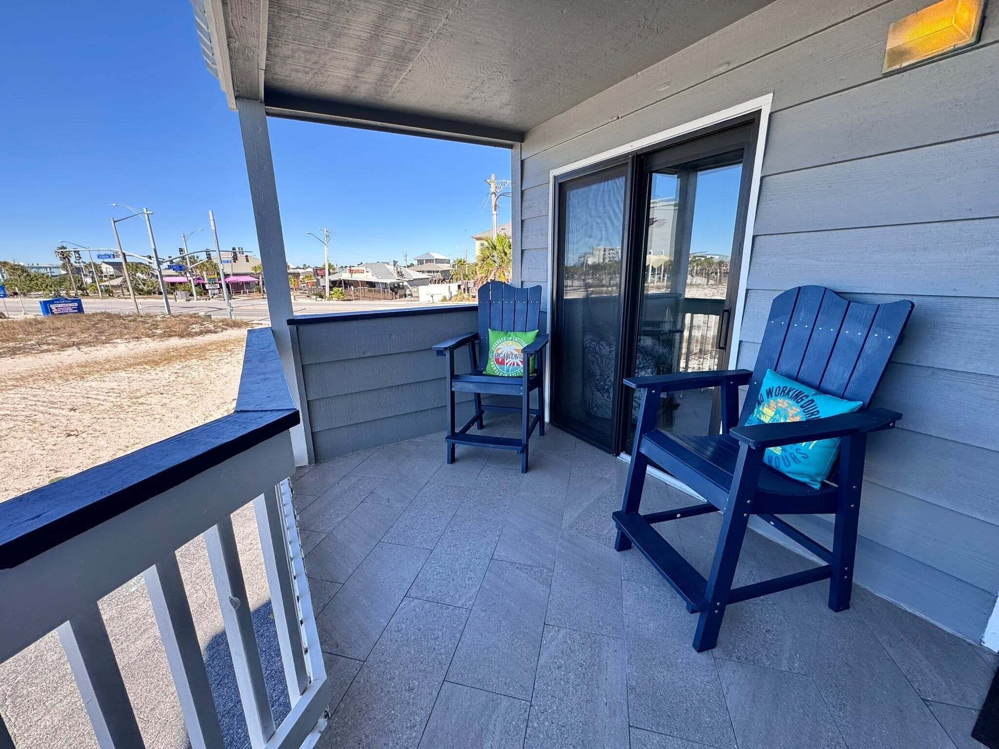 A covered balcony with two Adirondack chairs, a glass door, and a view of the beach and buildings.