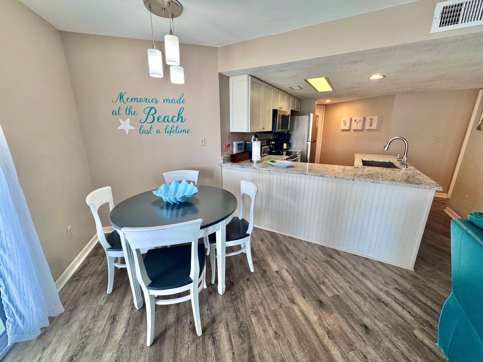 A compact kitchen with a dining area, featuring a kitchen island with a sink, a dining table with chairs, and a wall with a decorative phrase.