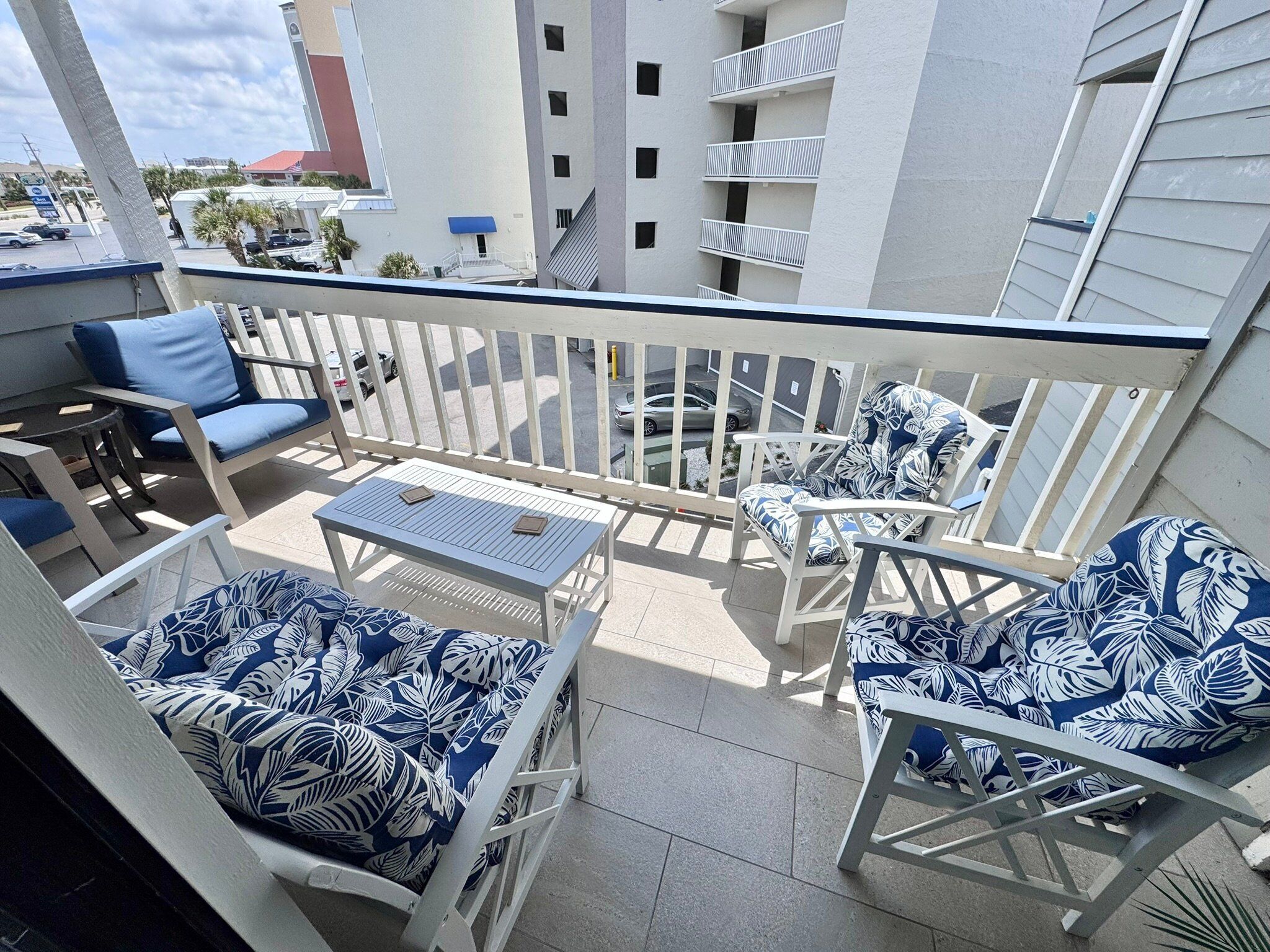 Un balcon doté de mobilier blanc, comprenant des chaises et une table, et offrant une vue sur un parking et des bâtiments en arrière-plan.