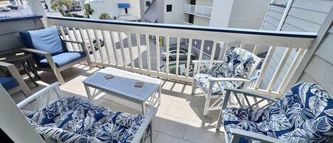A balcony with white furniture, including chairs and a table, and a view of a parking area and buildings in the background.